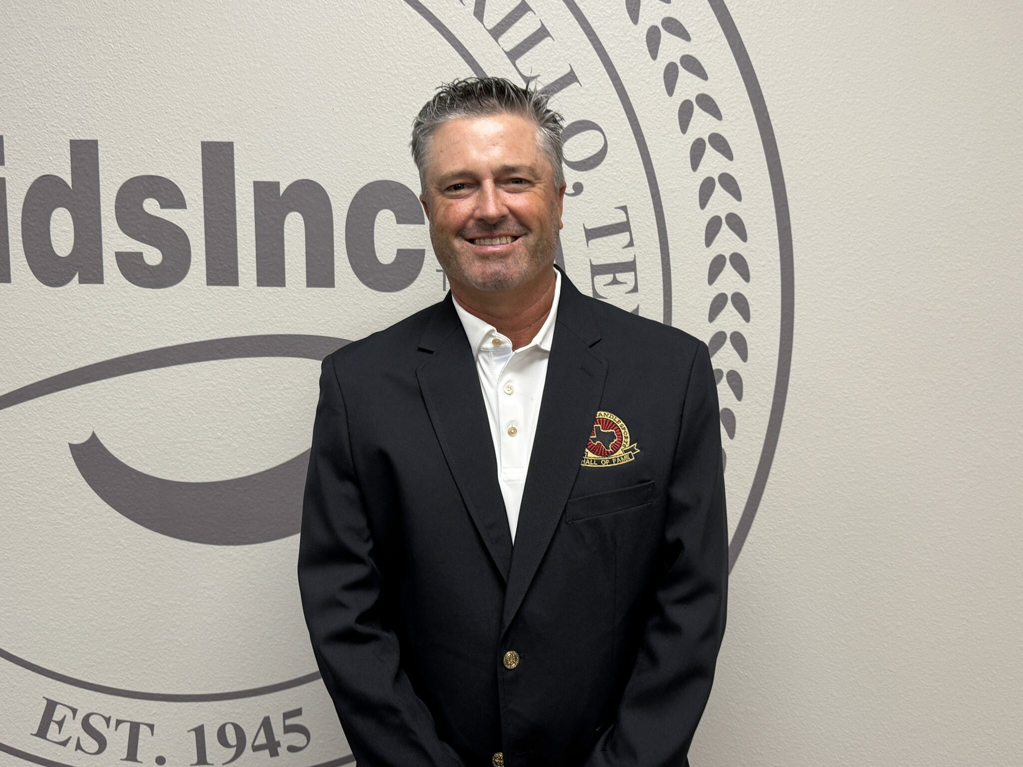 IMG_5296 | Press Pass Sports Smiling man in a dark suit stands in front of a large circular emblem on a beige wall.