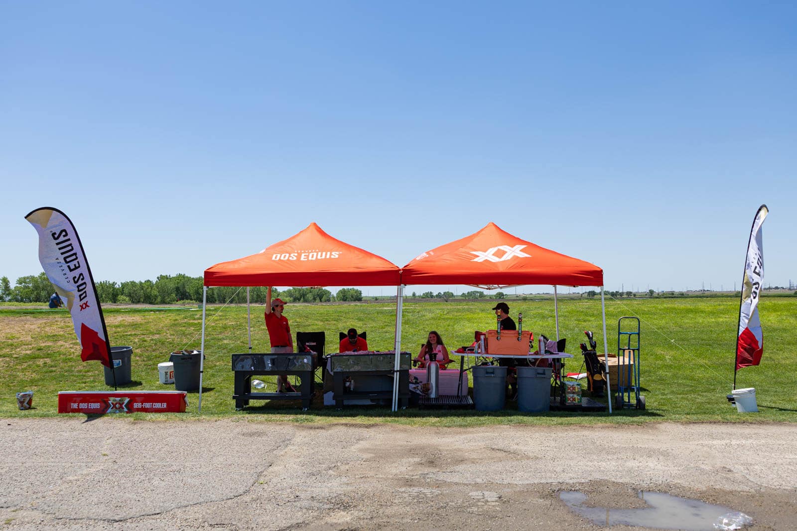 Promotional booth with orange canopies and people serving drinks at an outdoor event in a grassy field under a clear blue sky.