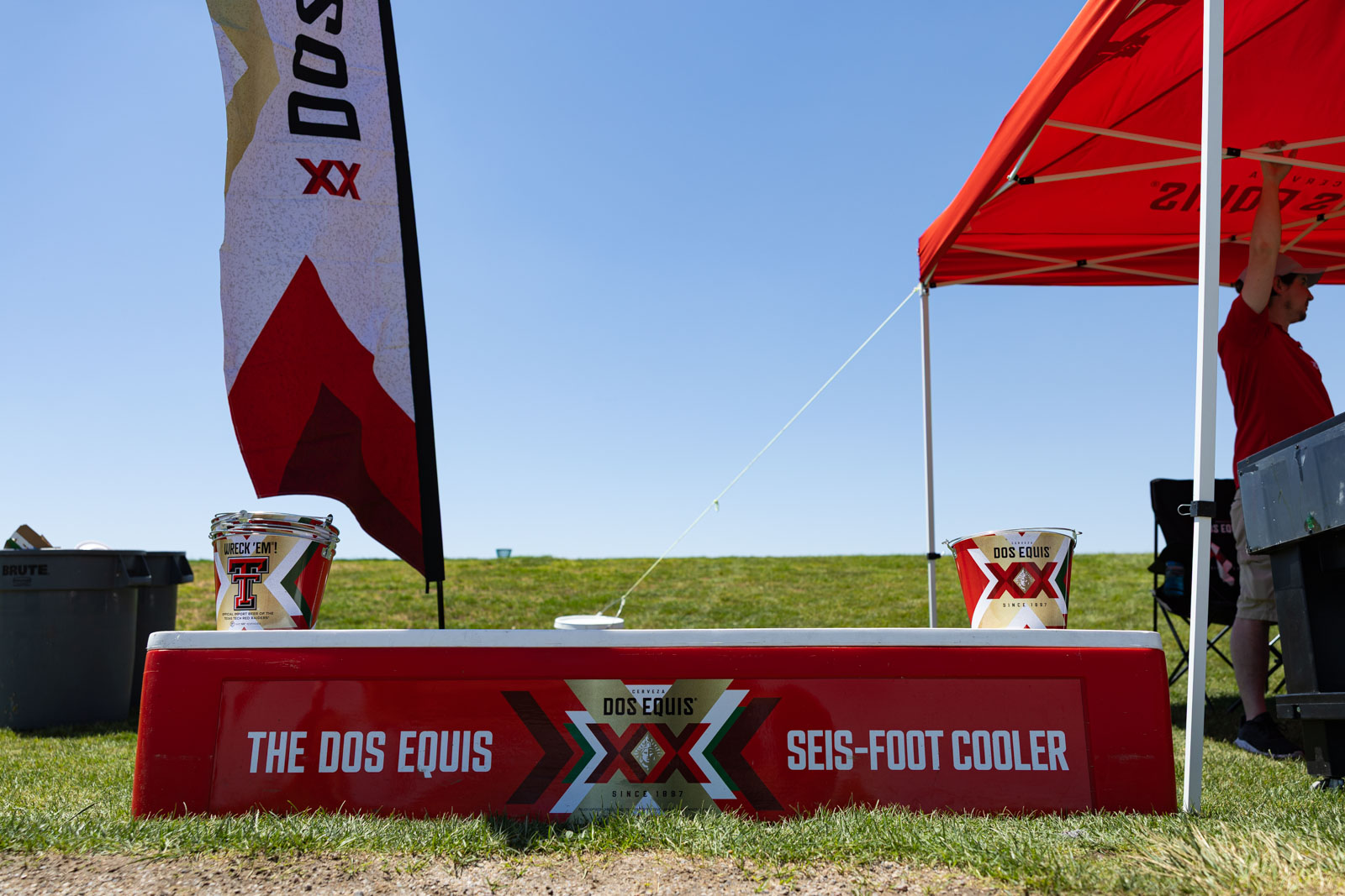 Red Dos Equis cooler on grass at an outdoor promotional booth with a bucket labeled Dos Equis and a red canopy in the background.