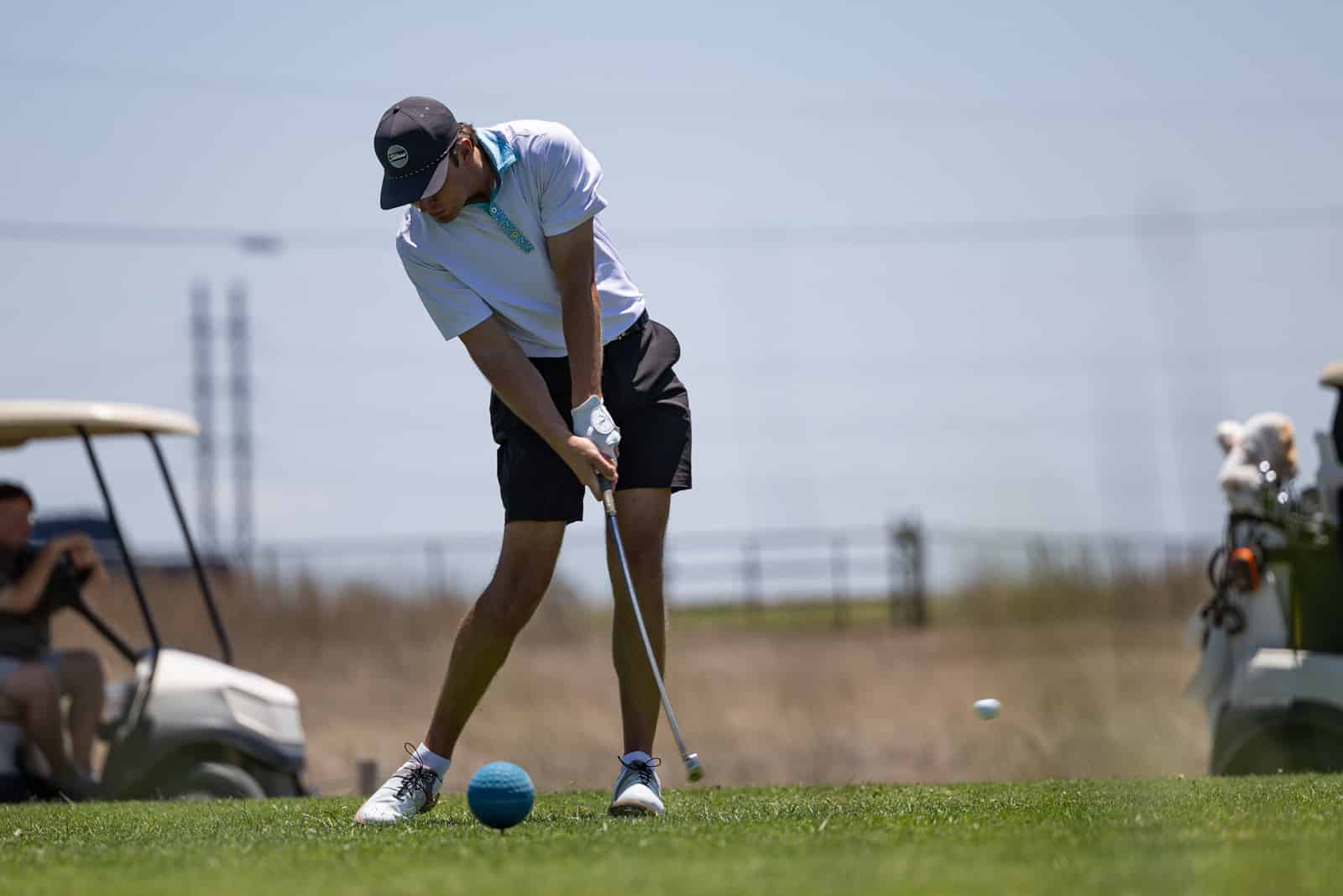 Male golfer in a white polo shirt and blue collar swings a club on a sunny golf course, with a golf cart and spectators in the background.