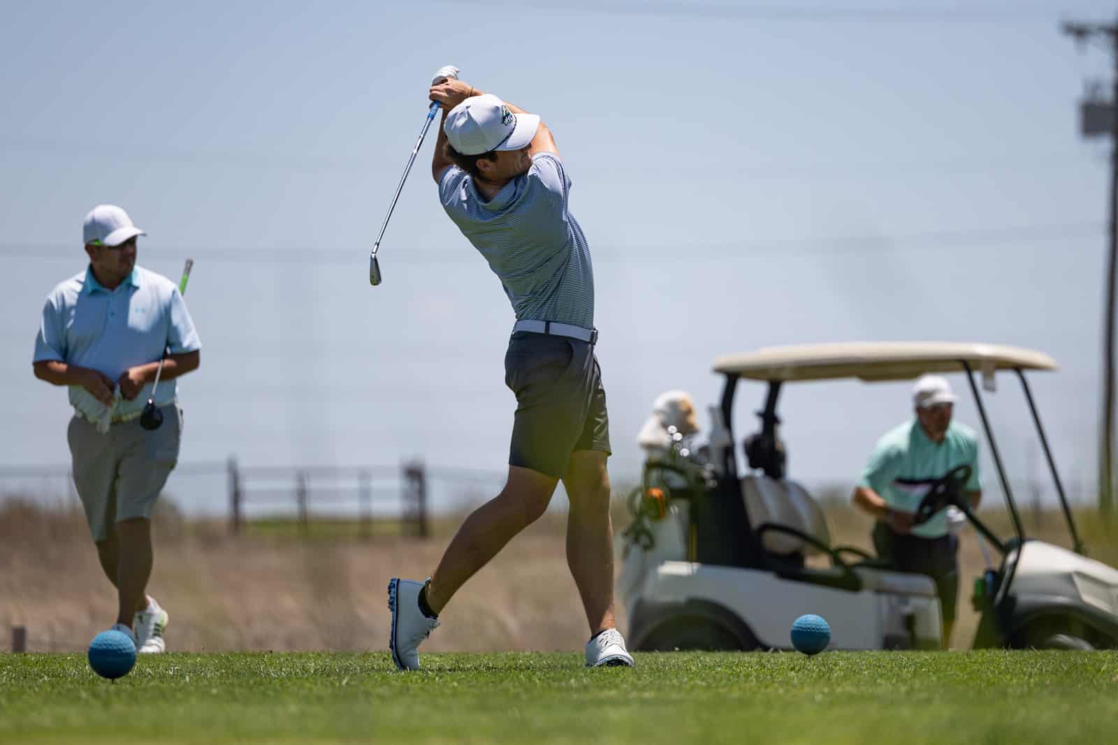 Golfer in a light blue shirt and white cap executing a full backswing on a sunny golf course, with a blue tee ball nearby.