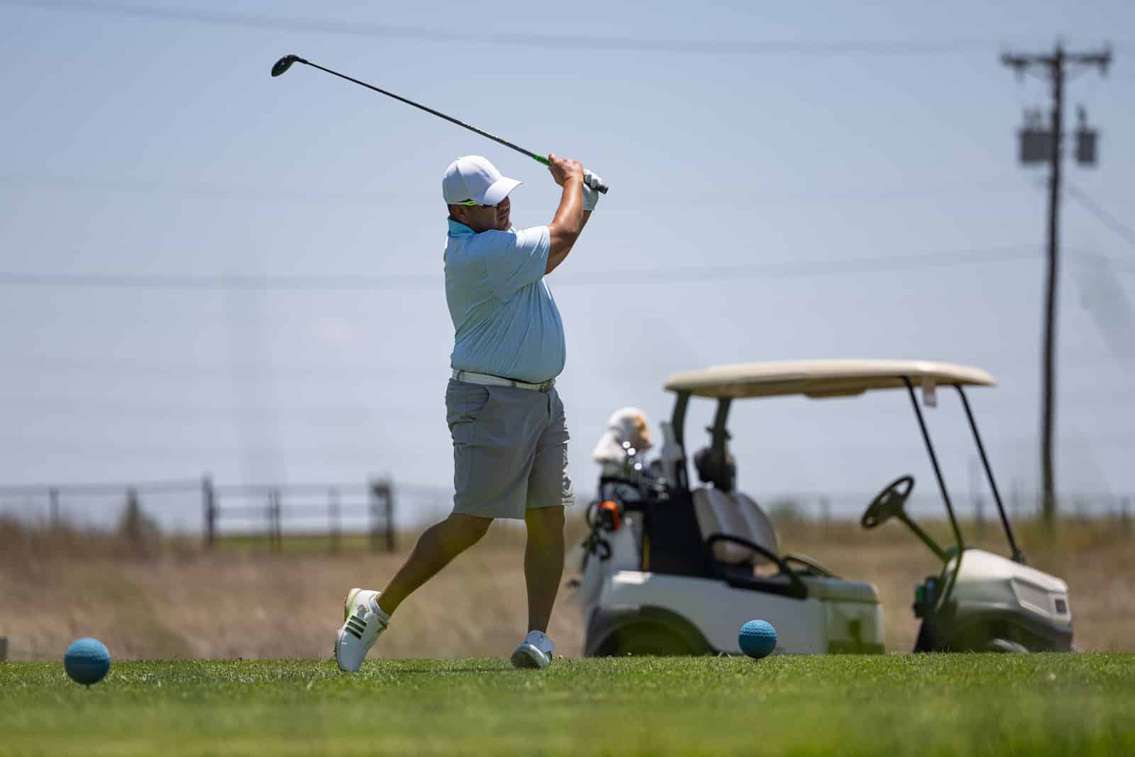 Golfer mid-swing on a sunny golf course, wearing a white cap and light blue polo; a golf cart is in the background.