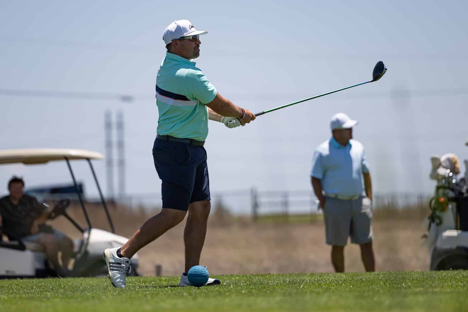 Male golfer in a teal shirt and white cap mid-swing on a sunny golf course, ball on tee in the foreground, spectators and a cart in the background.