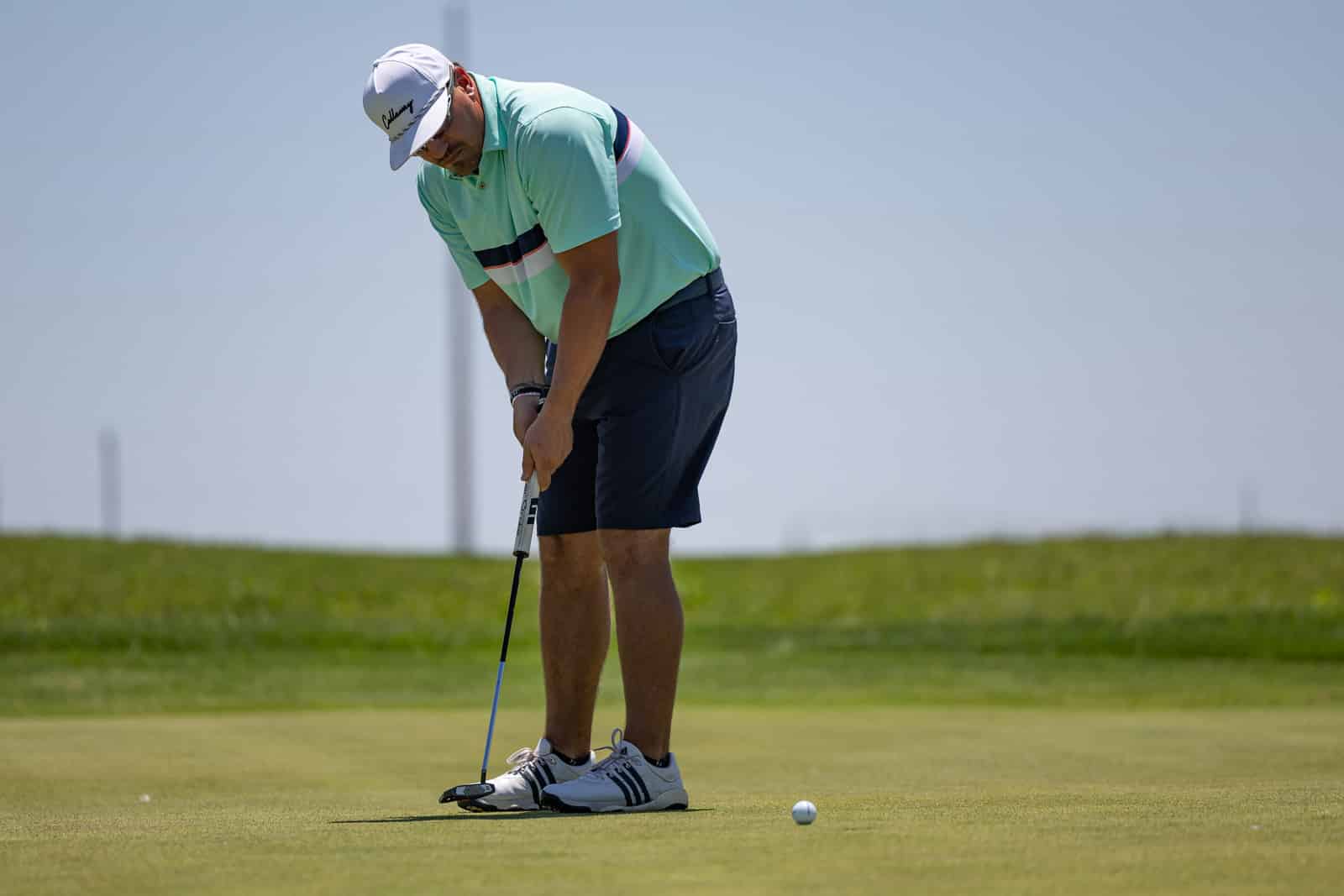 Golfer lining up a putt on a sunny green, wearing a mint polo and white cap.