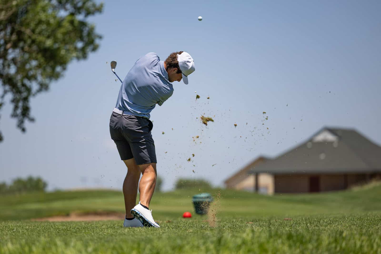 Golfer in a blue striped shirt and cap swipes a bunker shot, sand flying as the ball lifts into the blue sky.