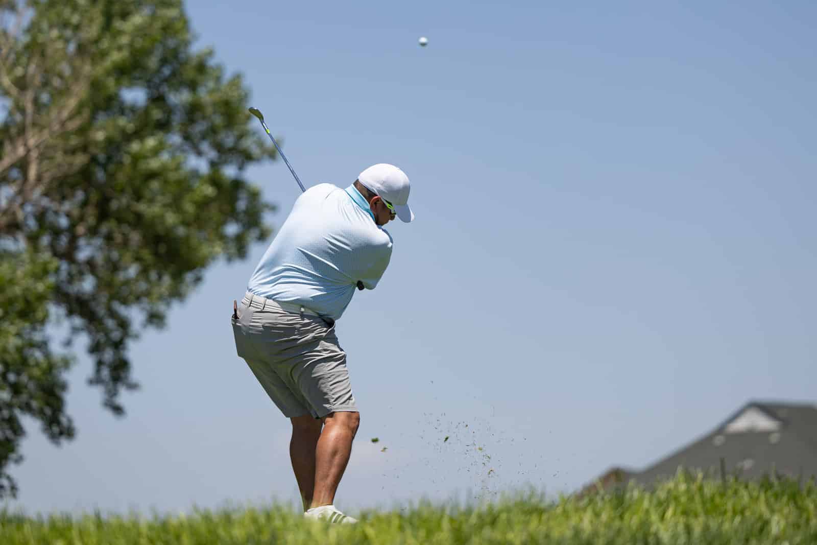 Golfer in a light blue shirt and white cap swinging a club with the ball in midair against a blue sky on a sunny golf course.