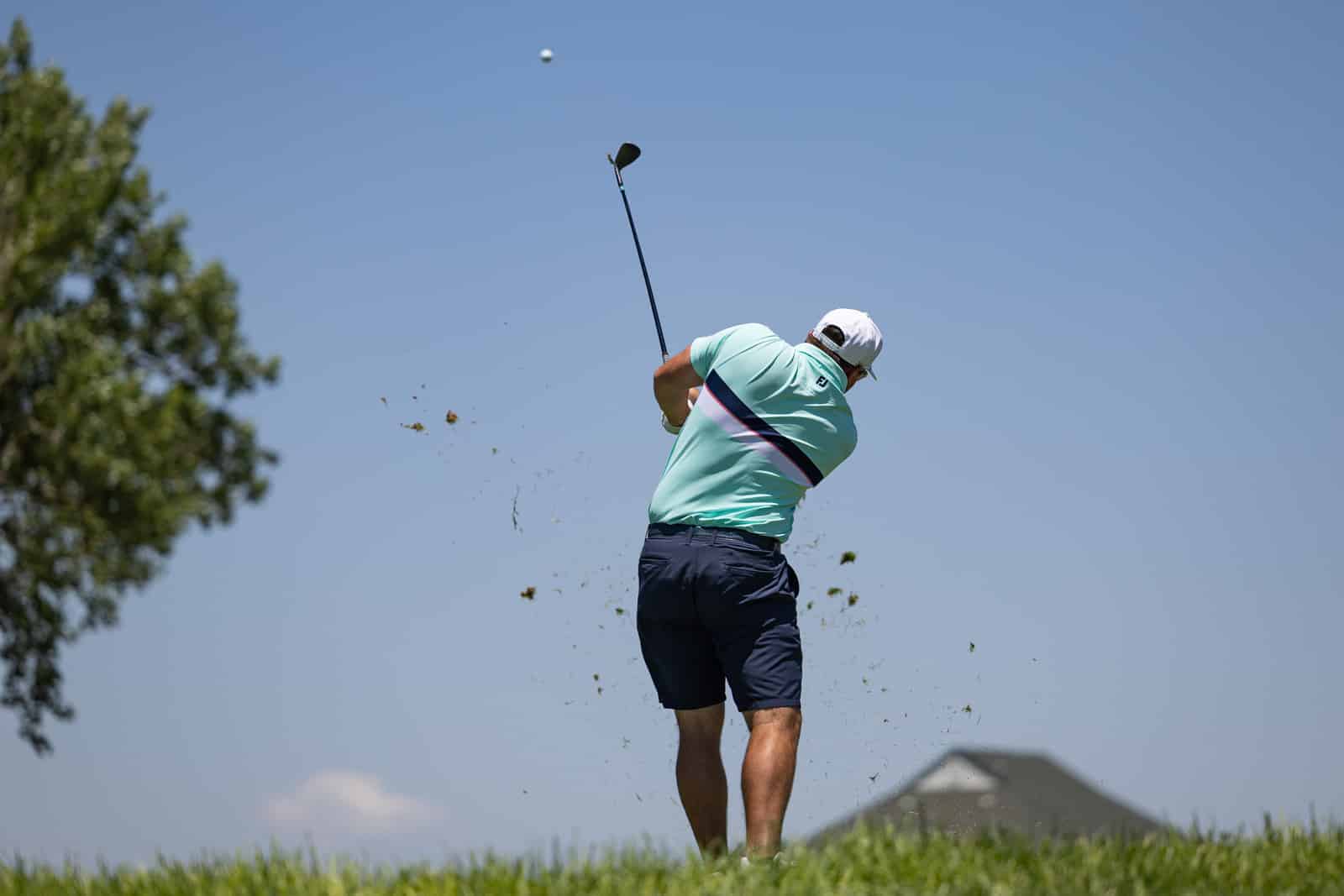 Golfer in a mint green shirt and navy shorts mid-swing on a sunny golf course as the ball rises.