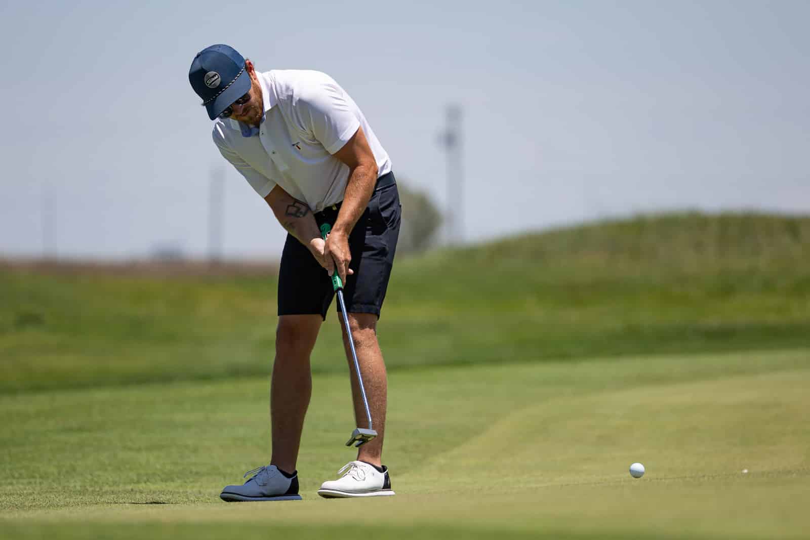 Golfer in a white polo and blue cap lining up a putt on a green, with a ball nearby