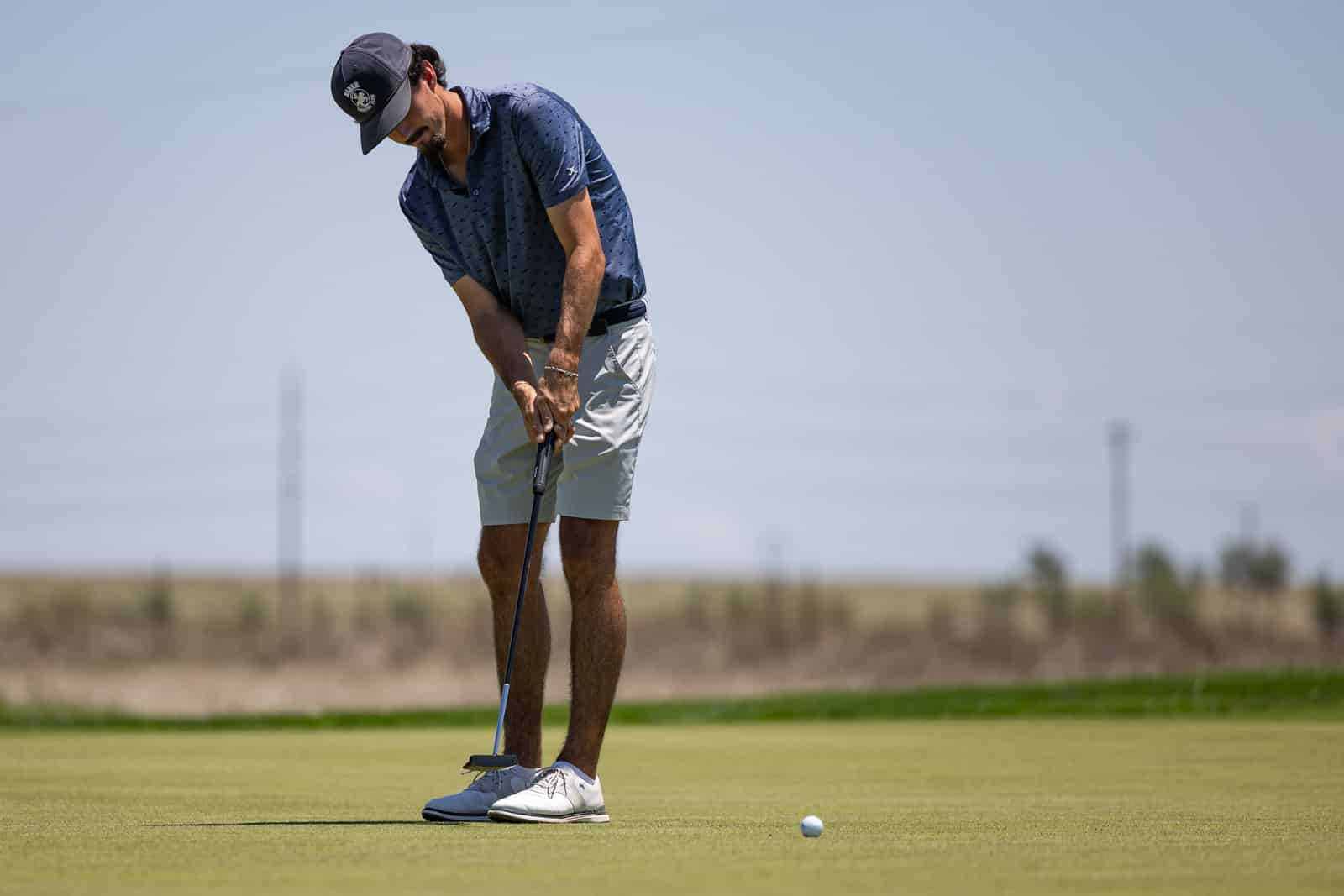 Golfer in a blue shirt and light shorts lining up a putt on a sunny golf green.