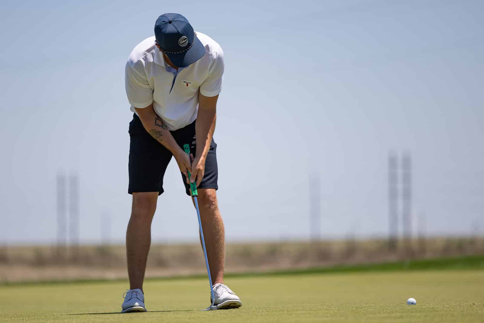 Golfer bent over, lining up a putt on a sunny golf course green with a ball nearby beneath a clear sky.