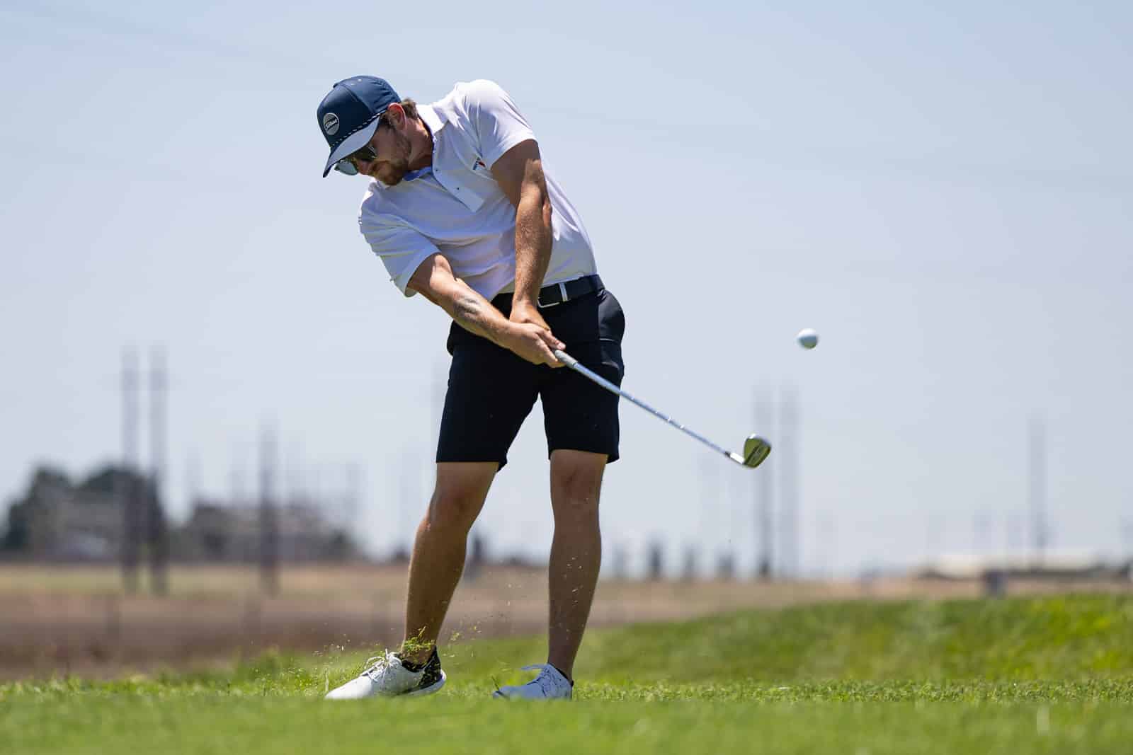 Golfer in white polo and blue cap mid-swing on a grassy course, ball airborne ahead of him.