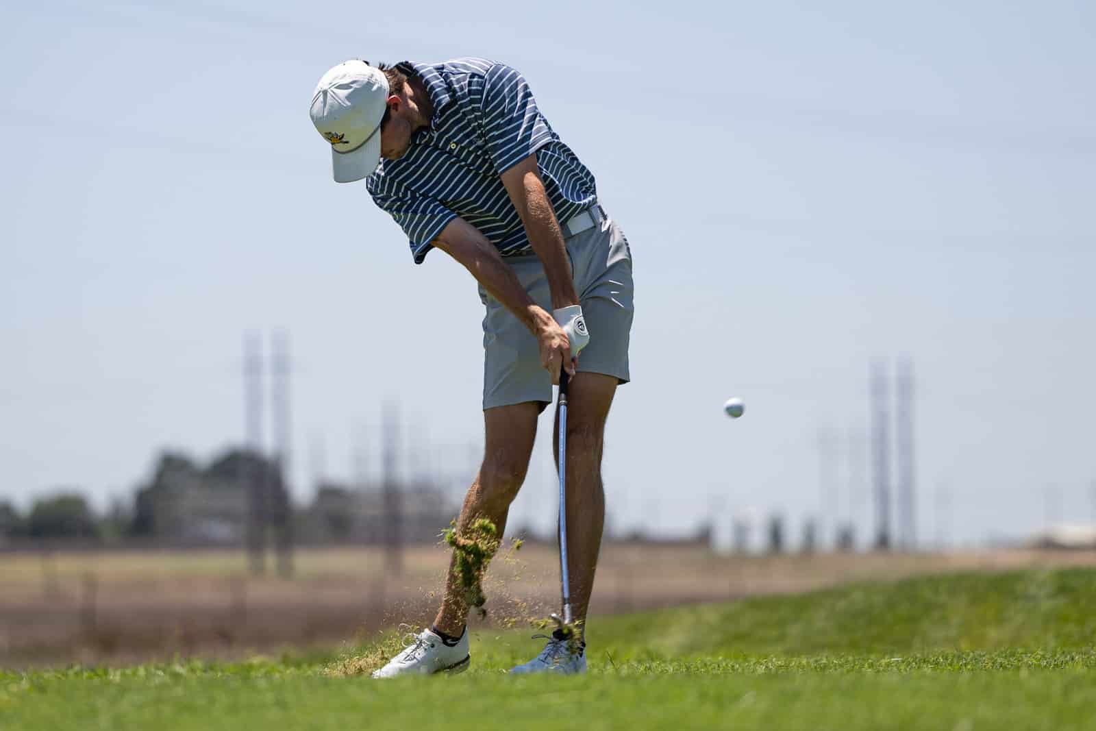 Golfer in a striped polo and cap swings a club, sending a ball into the air as dirt kicks up near his feet on a sunny course.