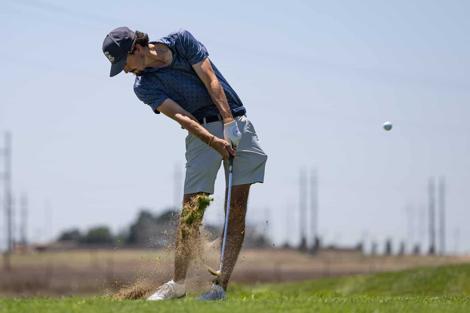 Golfer in a blue shirt and cap swipes a bunker shot, sand flying up from the sand trap as he follows through.