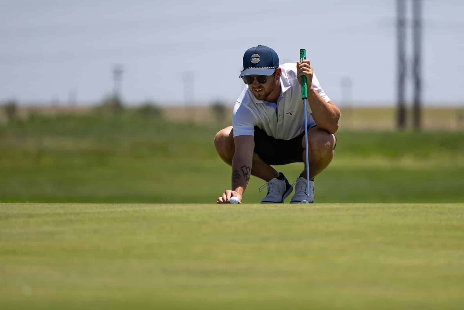 Golfer crouching on the green, lining up a putt with a blue-green putter nearby, wearing a white polo and navy cap with sunglasses.