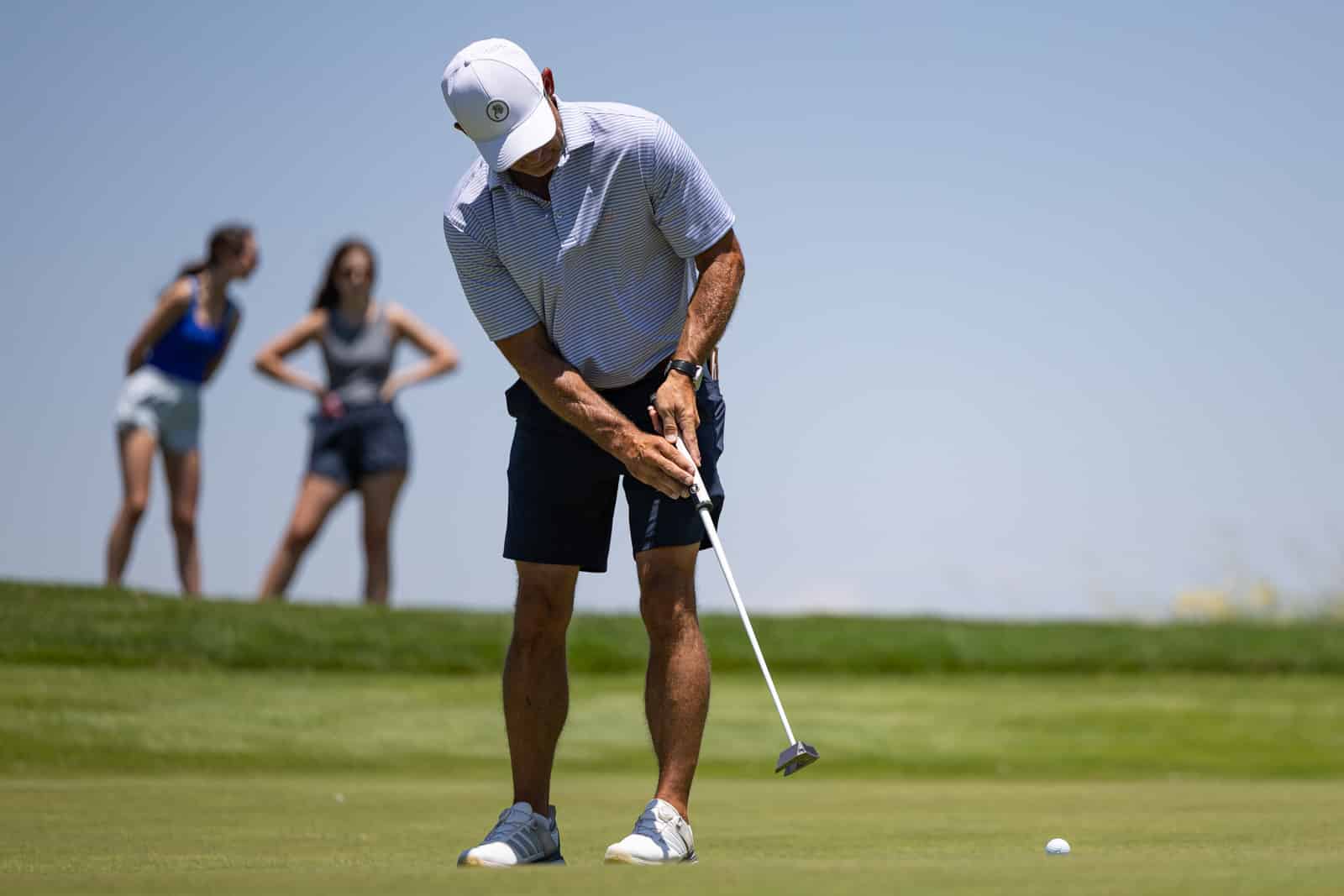 Male golfer lining up a putt on a sunny green, with two women watching in the background.