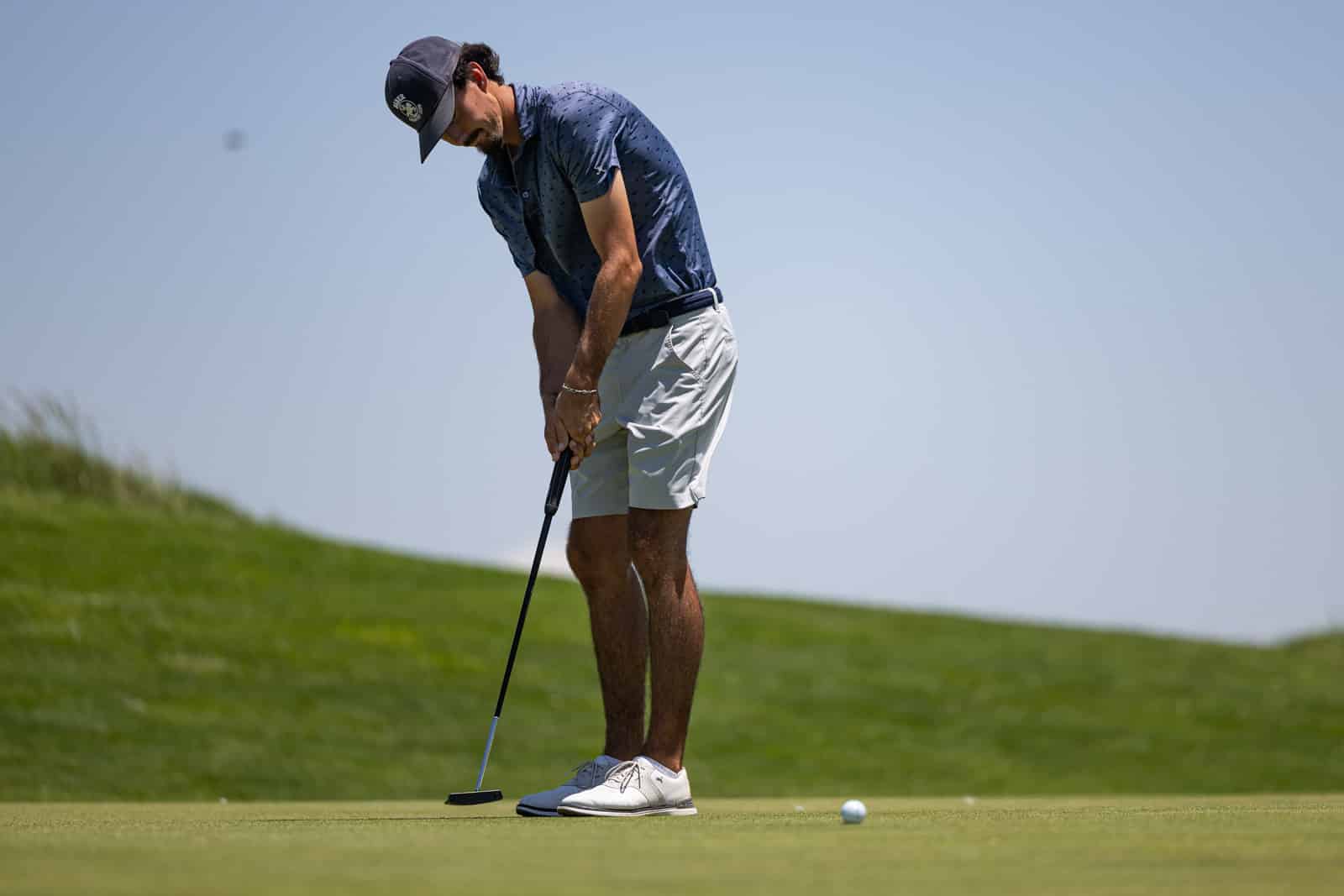 Golfer lining up a putt on a putting green with a blue polo shirt and white shorts.