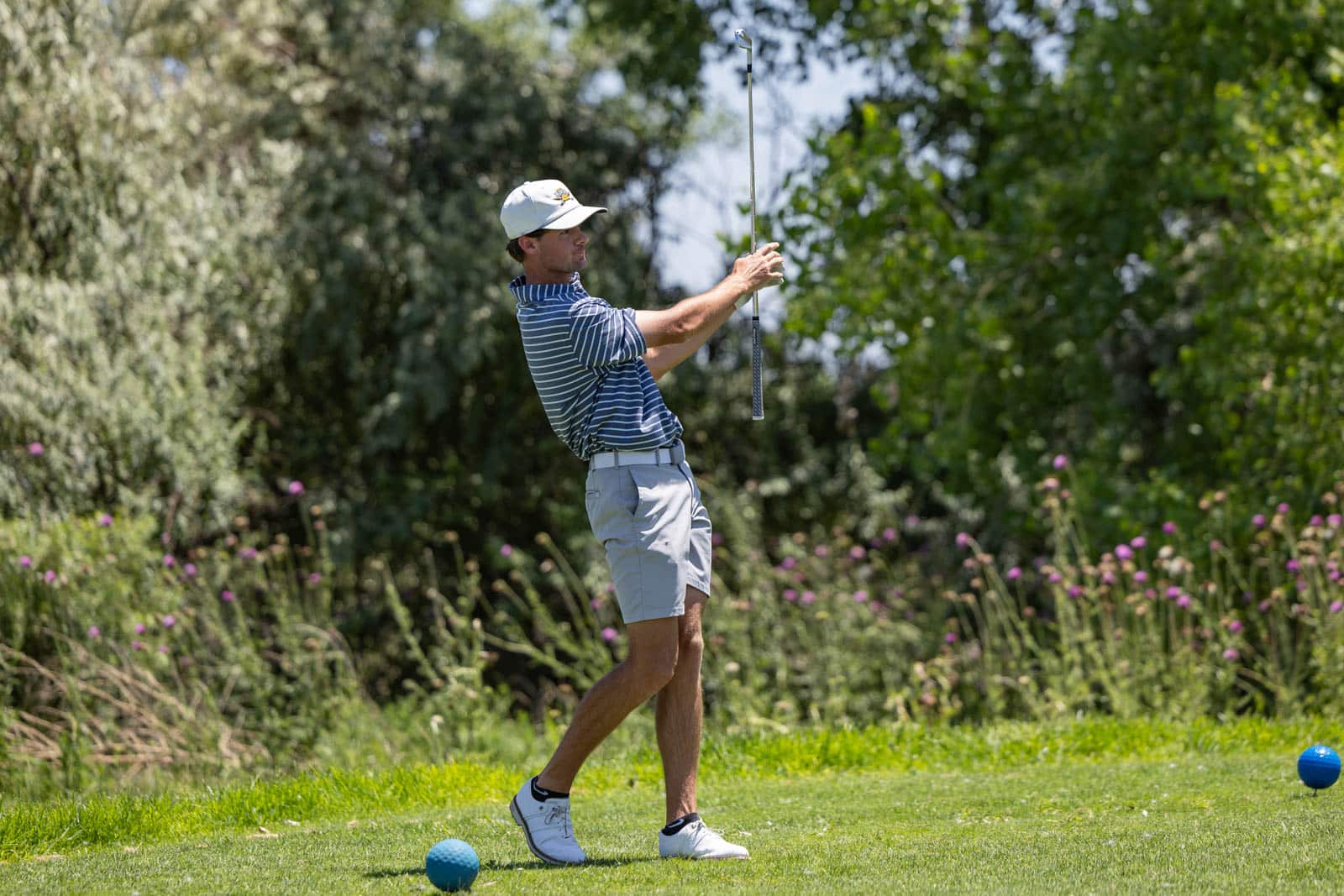Golfer in striped polo and white cap mid-swing on a sunny golf course with blue balls nearby.
