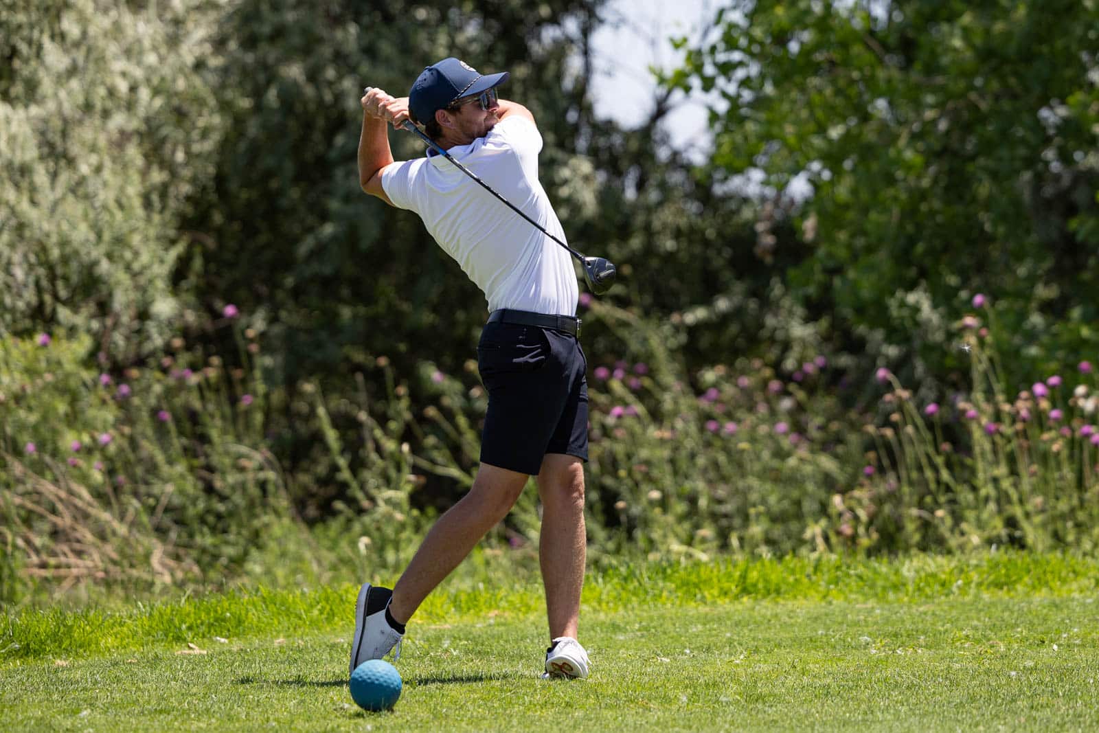 Golfer in white polo and navy cap swings a driver on a sunny course, ball teed up on a blue ball marker nearby