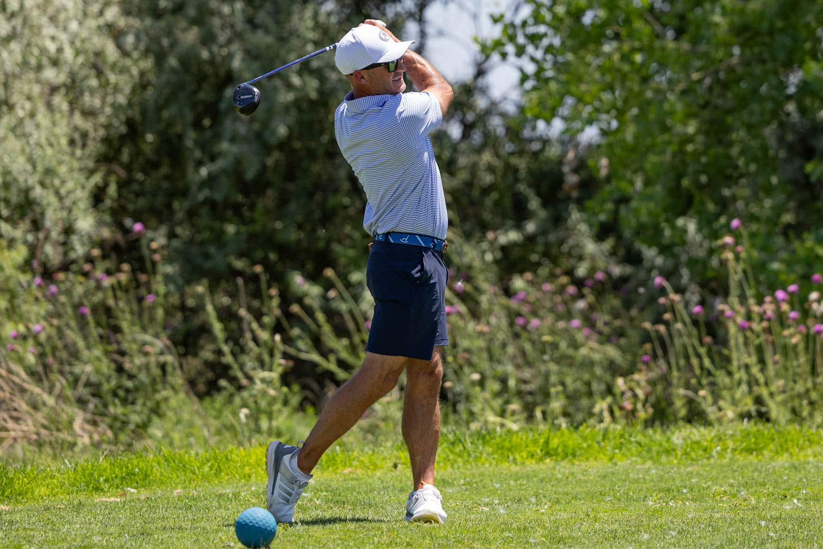 Golfer mid-swing on a sunny golf course, wearing a striped polo, navy shorts, and a white cap with a teal ball at his feet.