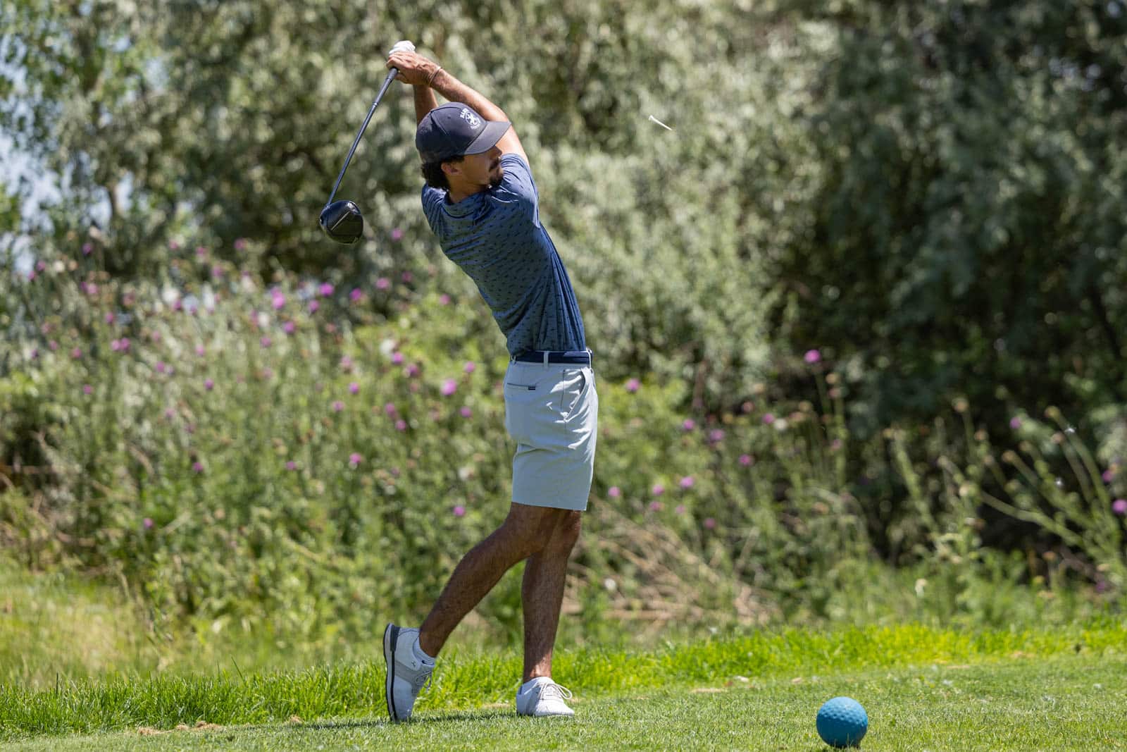 Male golfer in a blue shirt and light shorts mid-swing on a sunny golf course, with a blue golf ball on the tee nearby.