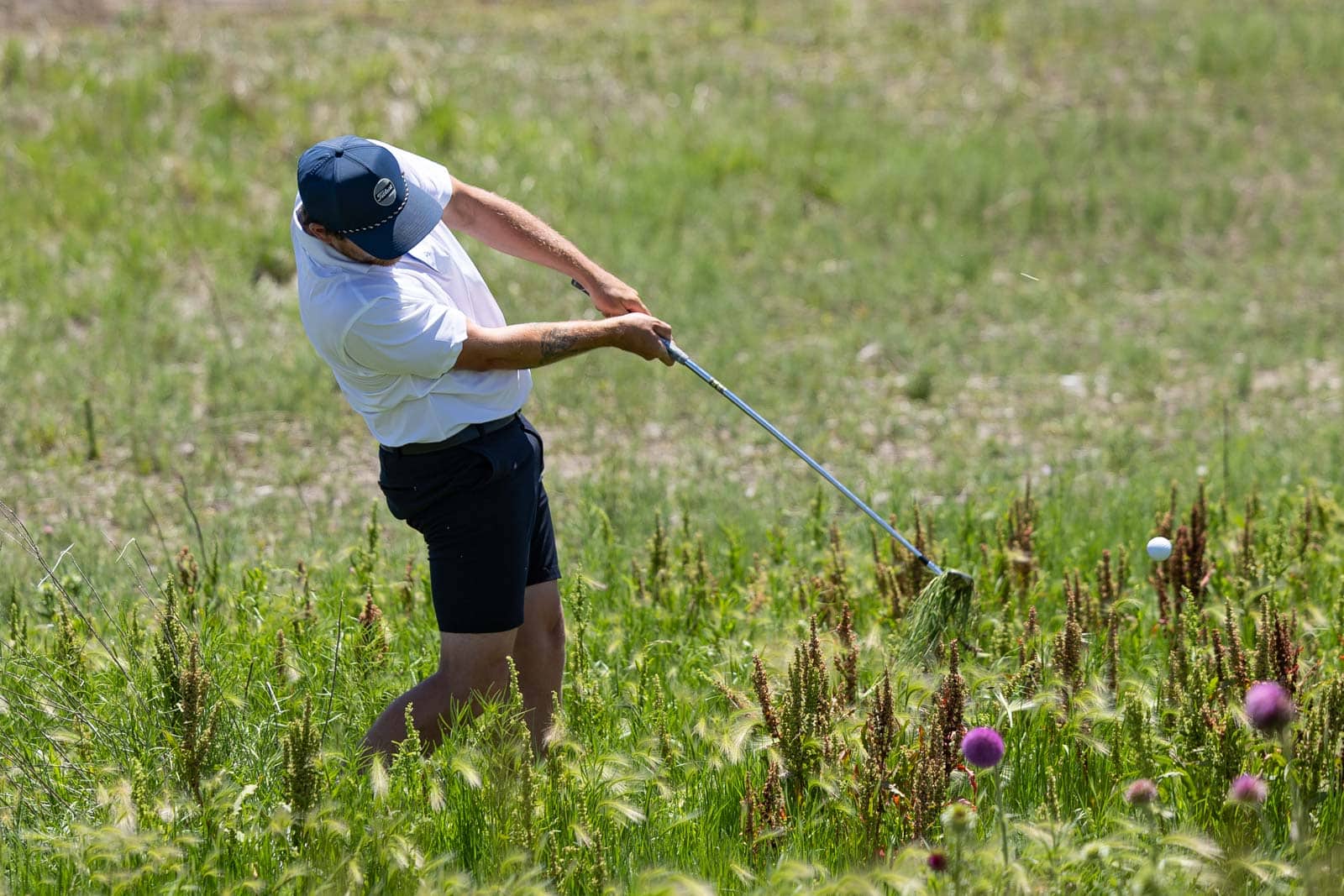 Man wearing a cap and white shirt swings a golf club in a tall grassy field, the ball just after impact.