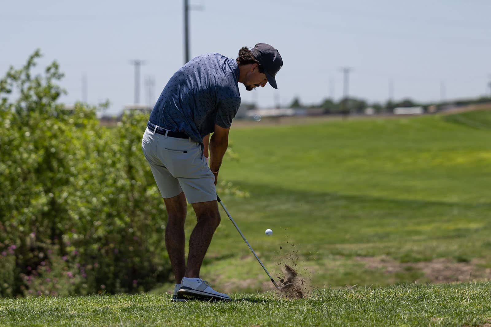 Golfer in a blue polo and light shorts executing a swing on a sunny golf course as a ball is struck from the tee area.