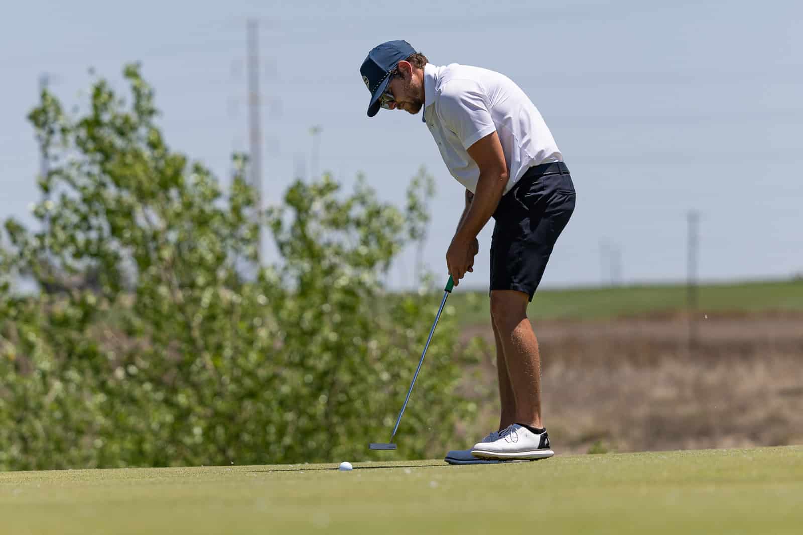 Golfer in white polo and dark shorts lining up a putt on a green golf course, wearing a blue cap and sunglasses.