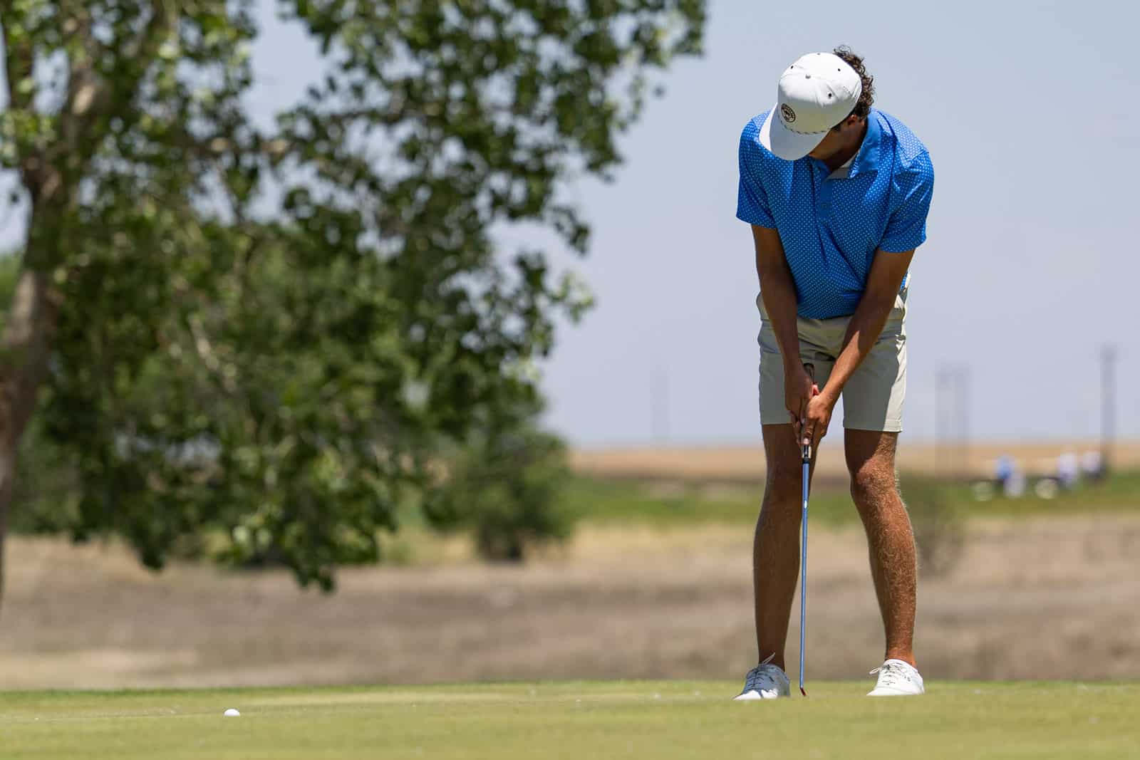 Male golfer in a blue polo and white cap lining up a putt on a sunny golf course.