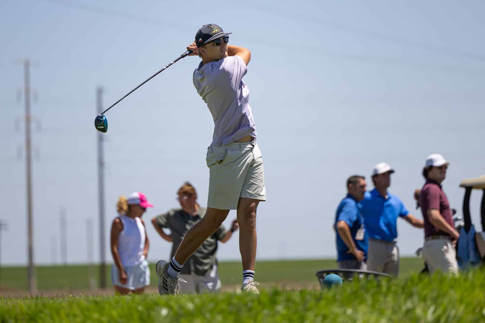 Golfer mid-swing on a sunny course, wearing a cap and sunglasses, with spectators in the background.