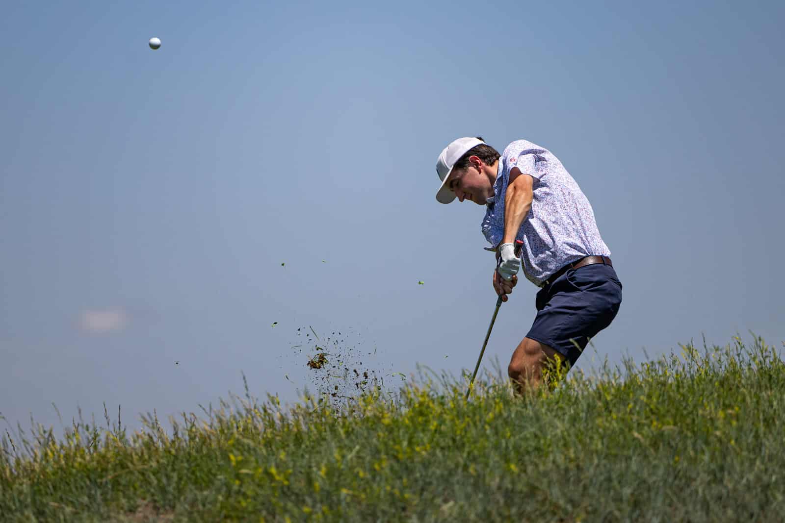 Golfer wearing a white cap and floral shirt swinging a club on a grassy hill as the ball flies away.