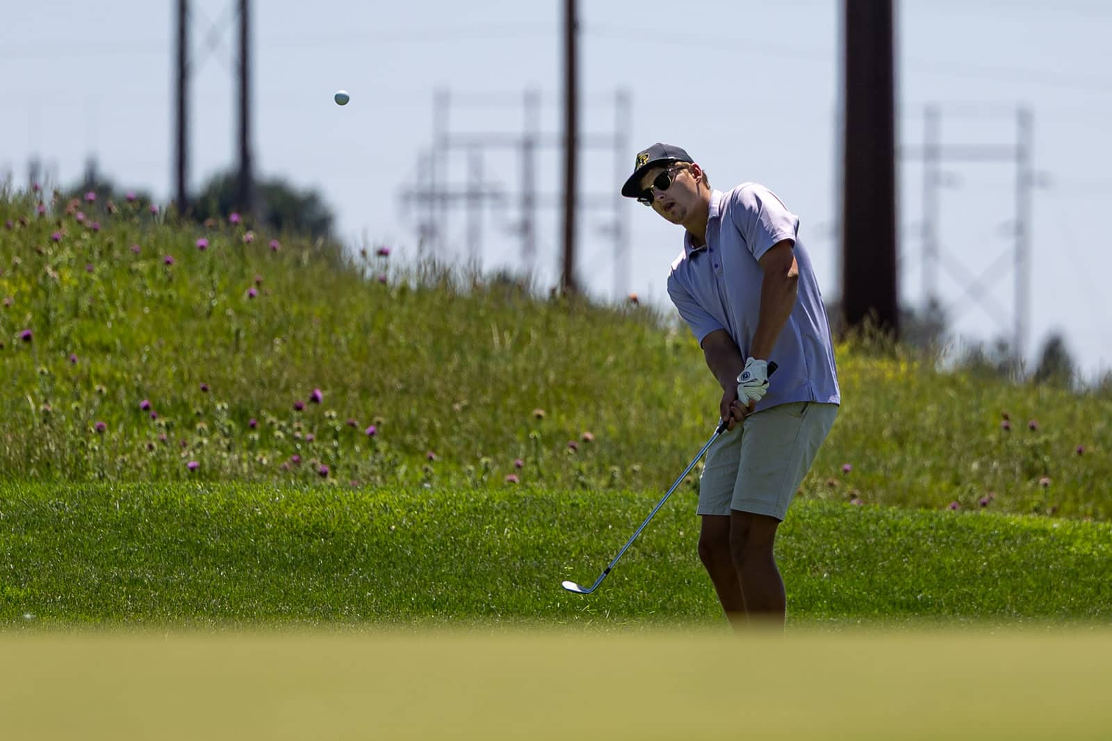 Man in a lavender polo and shorts swings a golf club on a sunny course, wearing a cap and sunglasses.