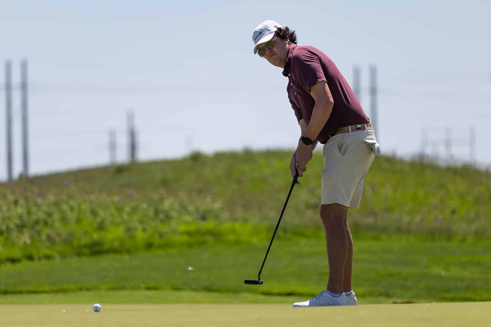 Man in maroon polo and white cap lining up a putt on a golf green, using a putter with a focused stance in bright outdoors setting