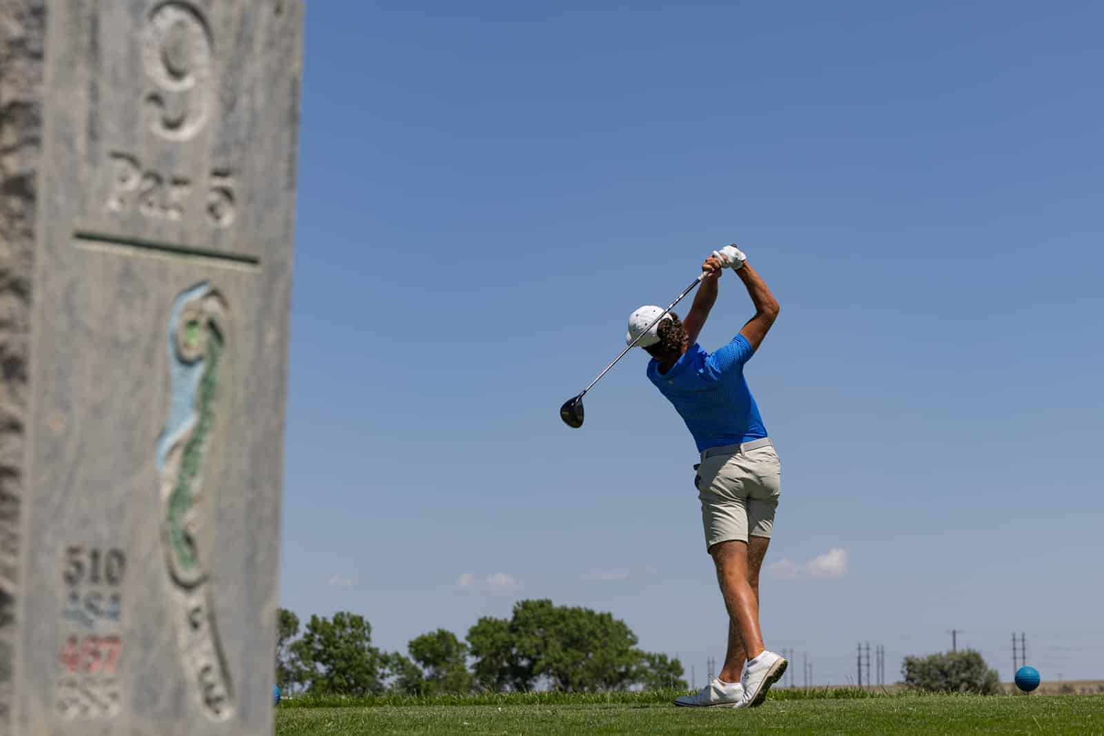Golfer in a blue shirt and white cap mid-swing on a sunny golf course, with a weathered stone marker in the foreground left.