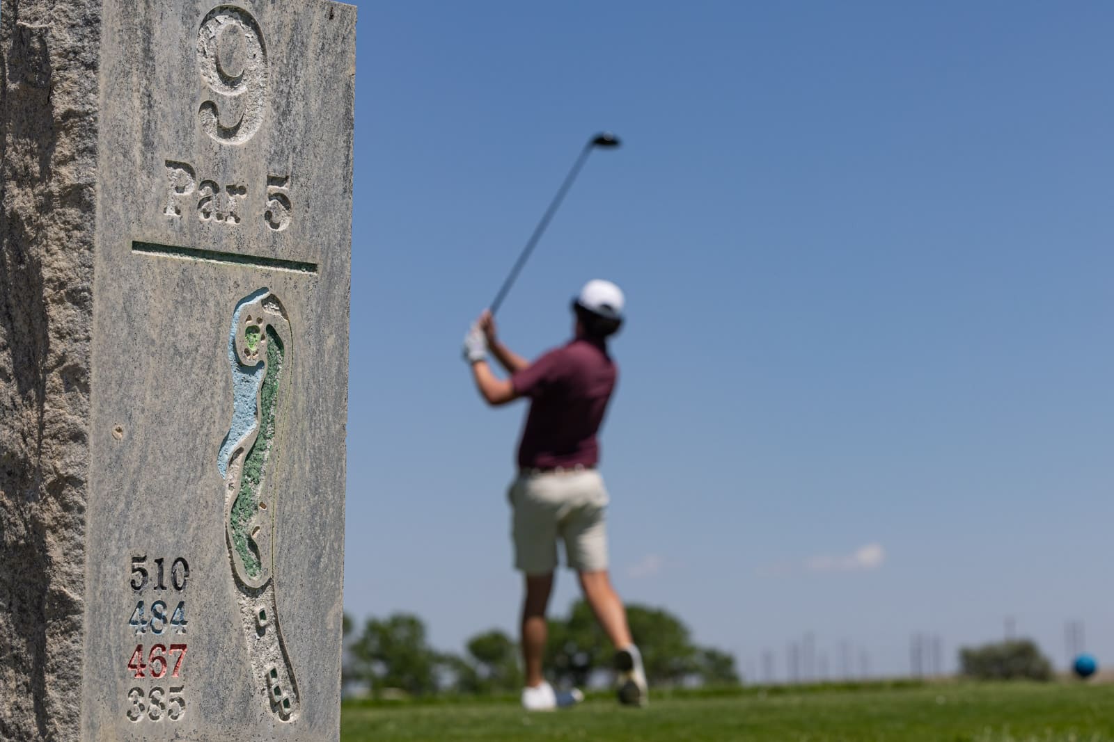 Stone golf-hole marker for hole 9, Par 5, with distances visible, while a golfer swings in the background on a sunny course.