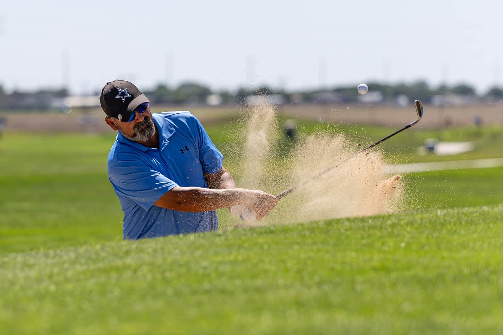 Male golfer in a blue polo swings from a sand bunker, sand spraying into the air as the ball pops up on a sunny course.