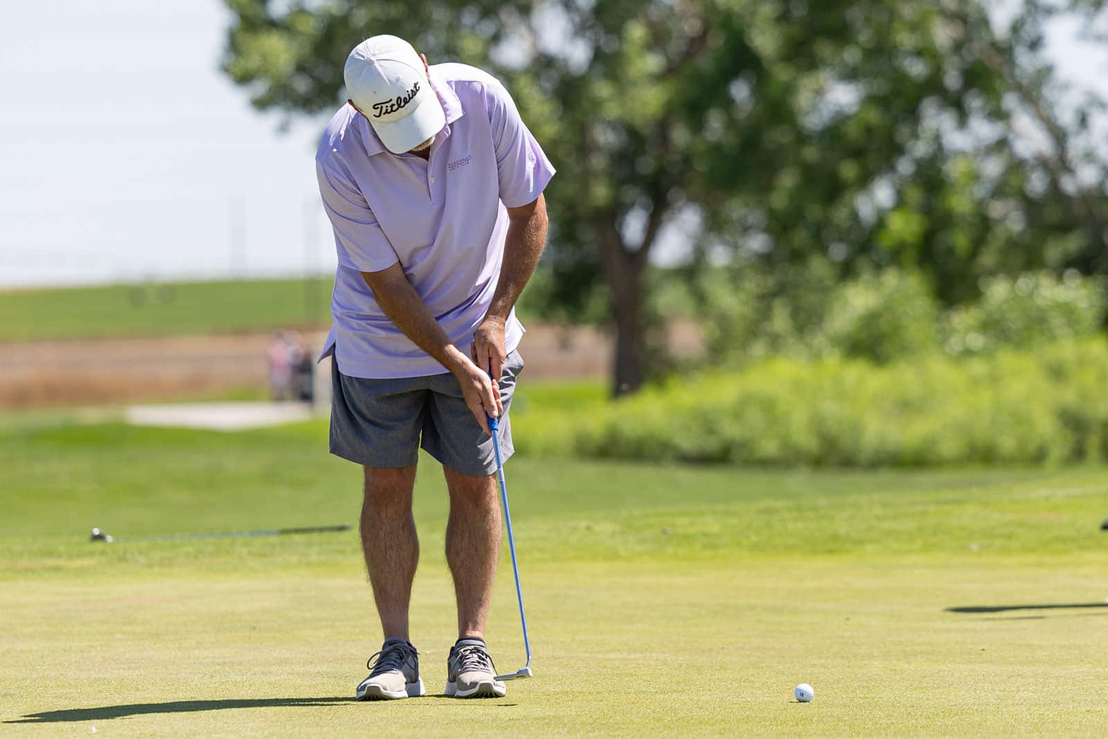 Man in a lavender polo and white cap lines up a putt on a golf green with a ball nearby