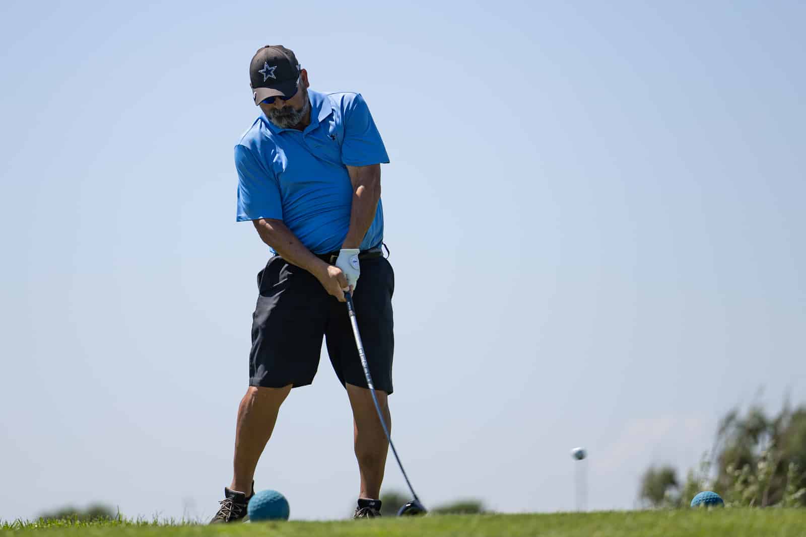 Golfer in a blue polo and dark cap tees off on a sunny golf course, hands on the driver.