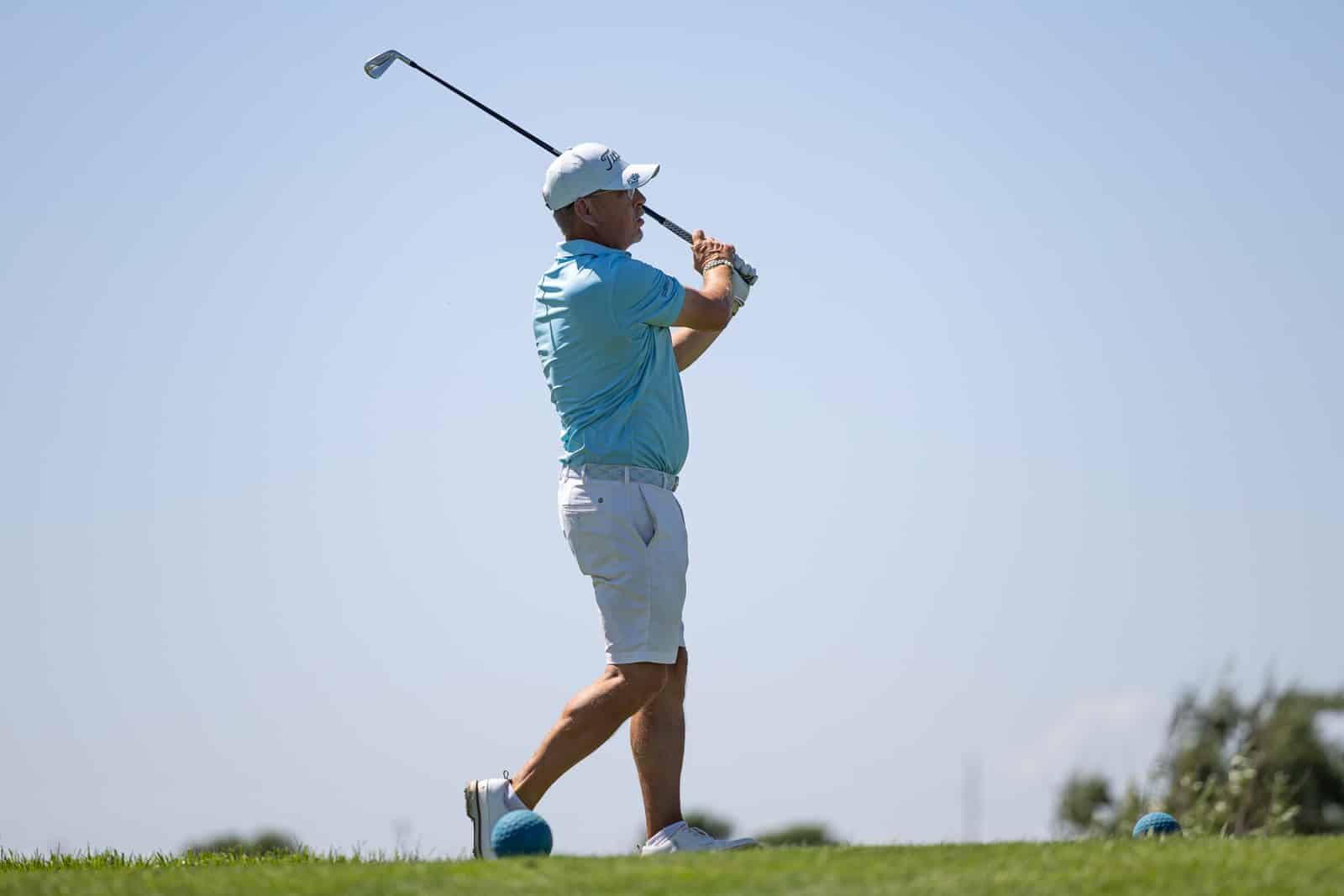 Male golfer in a light blue polo shirt and white shorts mid-swing on a sunny golf course with a blue golf ball at his feet.