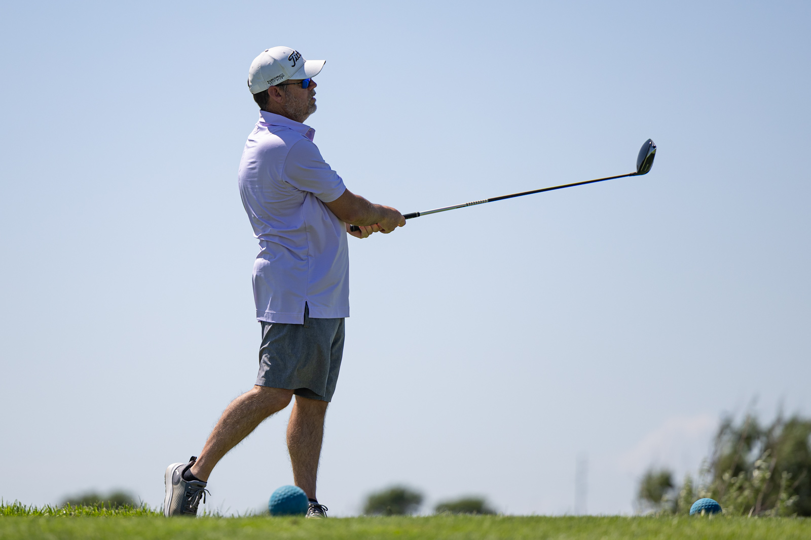 Man in a light purple polo and gray shorts mid-swing with a golf club on a sunny golf course.