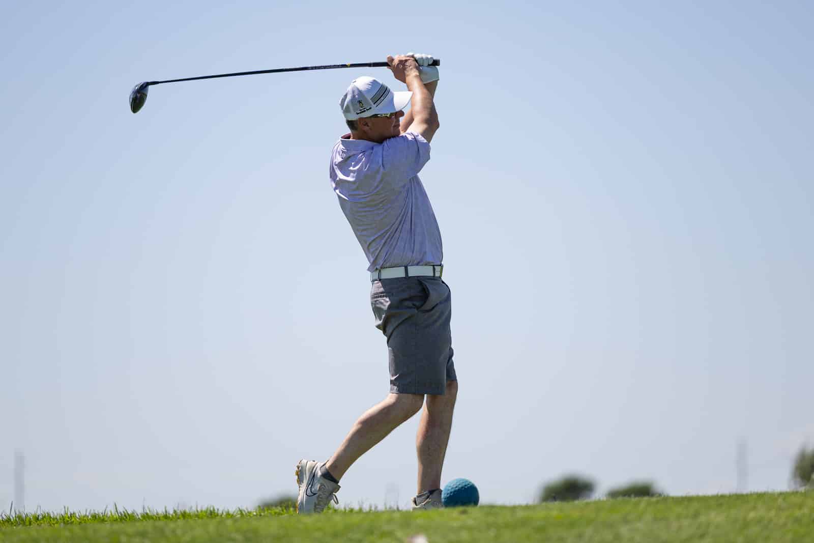 Golfer in a light purple shirt and gray shorts mid-swing on a sunny golf course, wearing a white cap and glove.