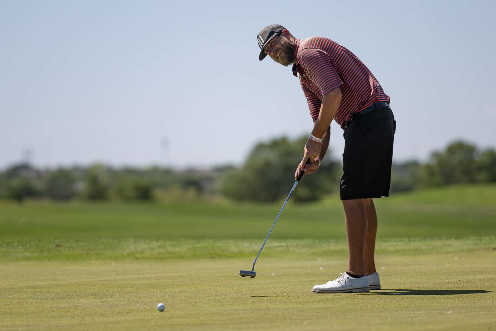 Man in a red-and-navy striped polo, black shorts, and a cap lines up a putt on a golf green under a clear blue sky.