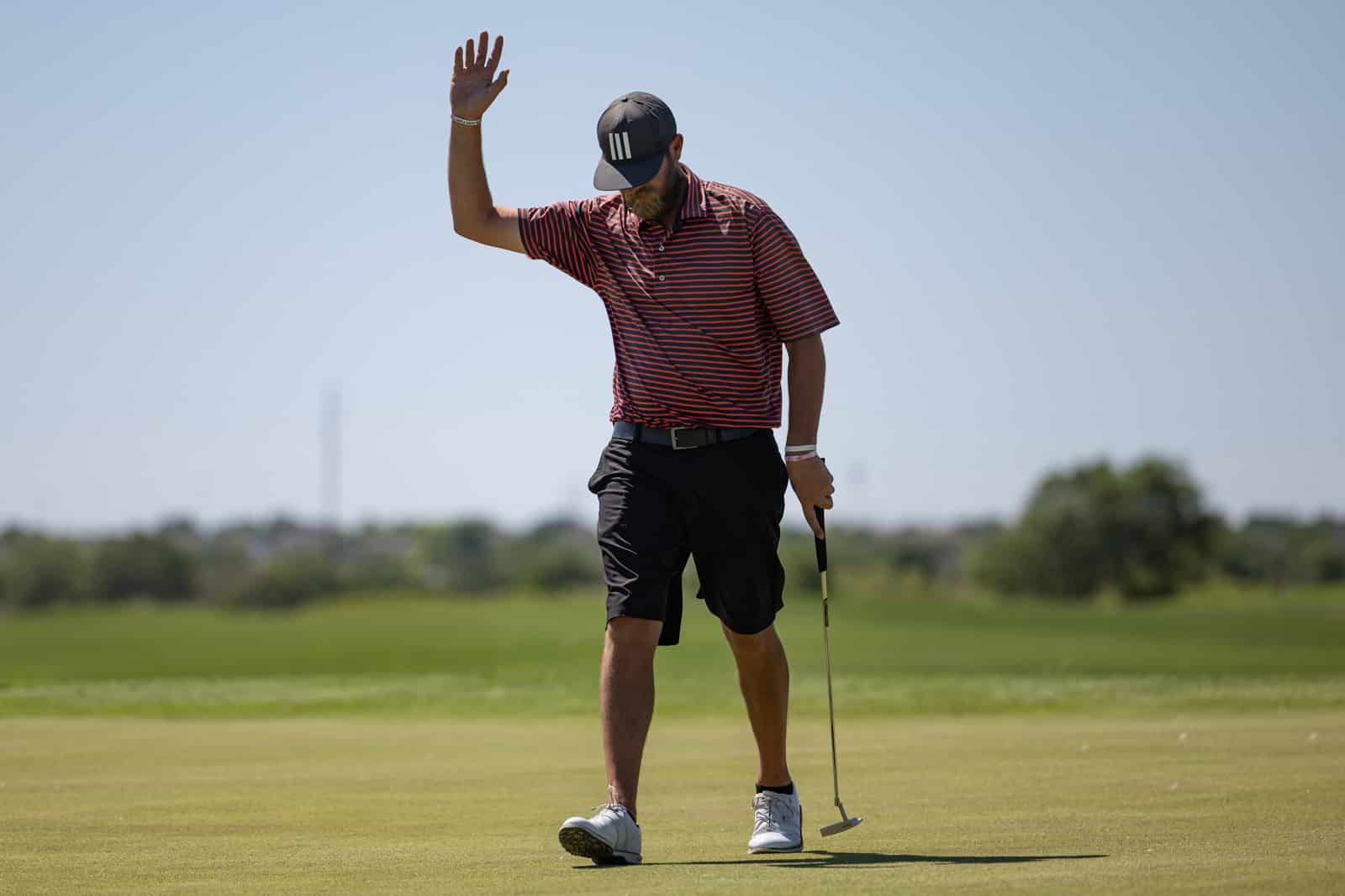 Golfer wearing a striped polo raises his right hand on a sunny golf green, holding a putter in his left hand.