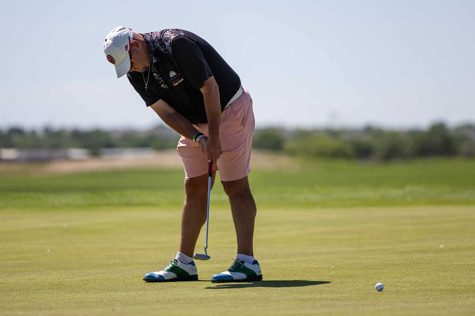 A man lines up a golf putt on the green, wearing a white cap, black shirt, and pink shorts, with a ball nearby.