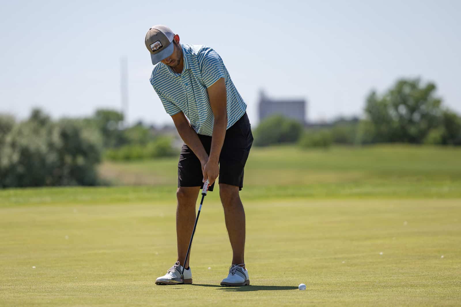 Man wearing a blue striped shirt and cap lines up a putt on a sunny golf green.