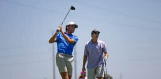 Golfer in a blue shirt mid-swing on a sunny golf course, with a teammate watching nearby.
