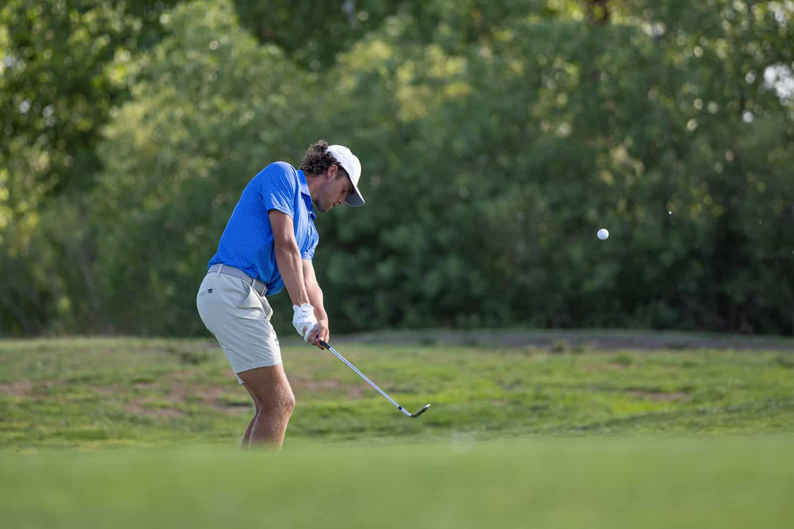 DosXX_Golf-74 | Press Pass Sports Golfer in blue shirt and white cap follows through on a golf swing as the ball shoots to the right.