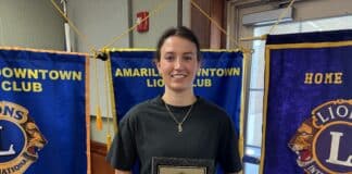 Young woman standing in front of Amarillo Downtown Lions Club banners, holding a framed award plaque.