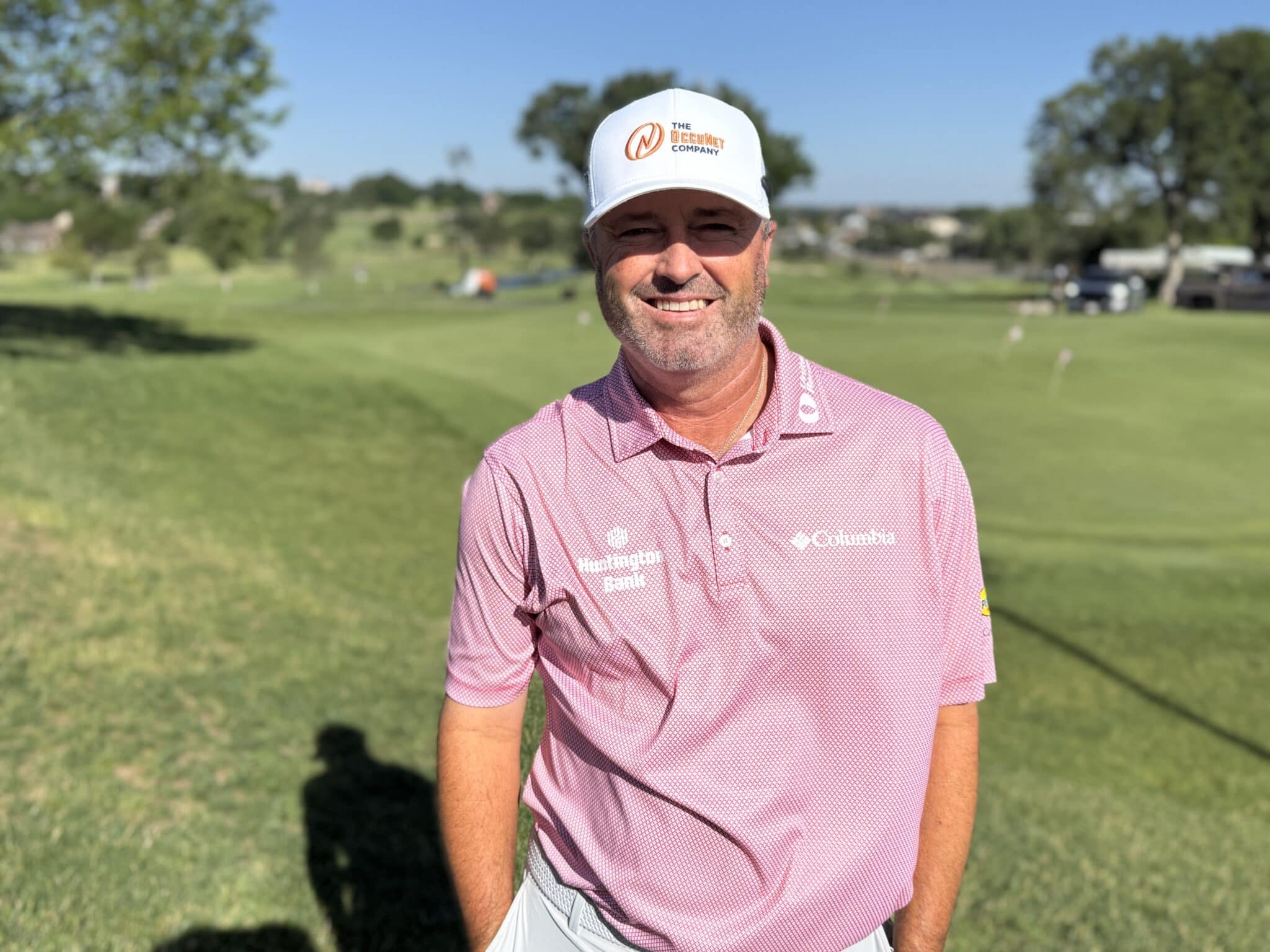 IMG_5341 | Press Pass Sports Man smiling at the camera on a sunny golf course, wearing a white cap and pink polo shirt.