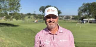 Man smiling at the camera on a sunny golf course, wearing a white cap and pink polo shirt.