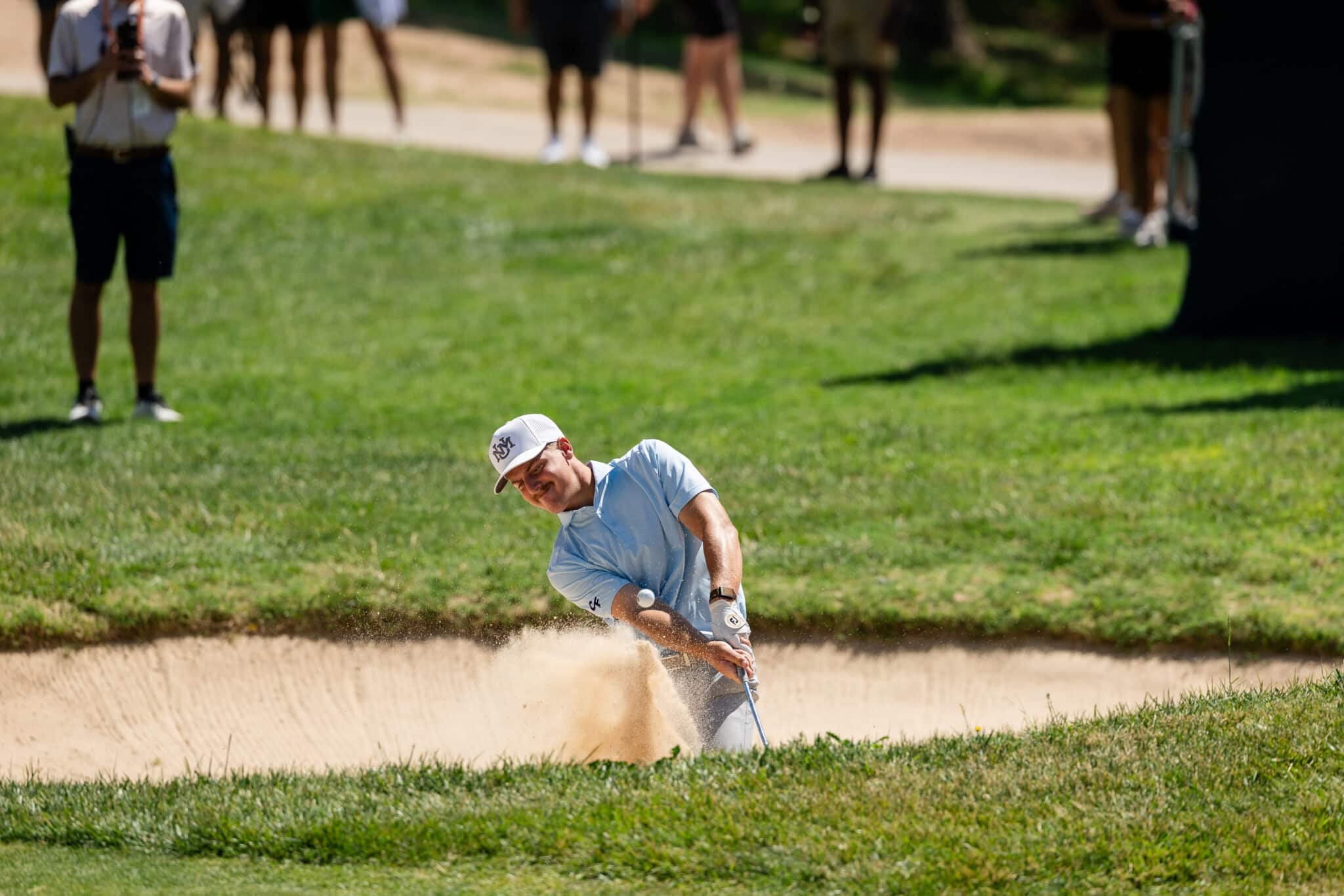 korn-ferry-occunet-classic-day-1-jpegJoe-159 | Press Pass Sports Golfer wearing a light blue shirt and white cap explodes from a sand bunker while spectators watch from the grassy edge (blurry background).